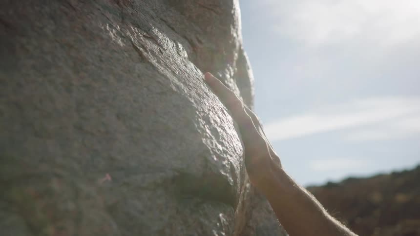 A person's hand gently glides across the sunlit surface of a large rock in slow motion, connecting with nature