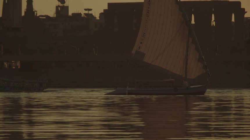 A traditional felucca sailboat glides along the tranquil Nile River at golden hour with the Luxor Temple silhouetted in the background