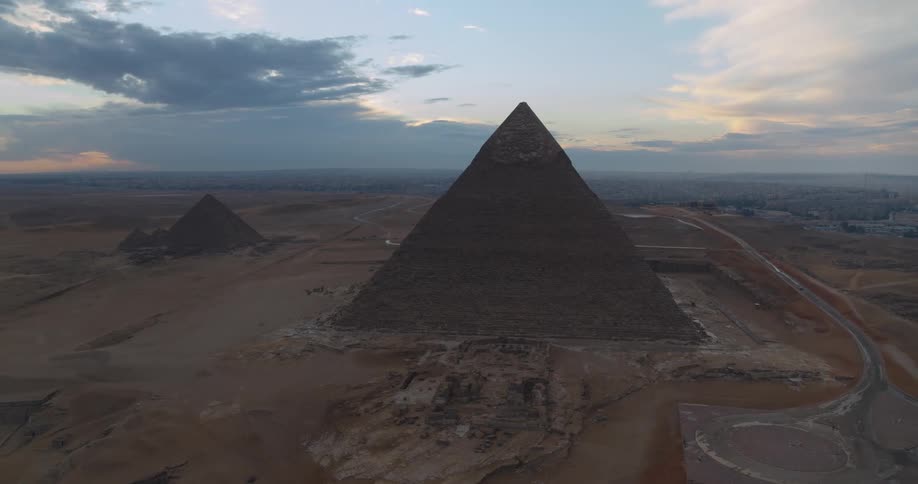 A slow aerial pullback shot reveals the majestic Pyramids of Giza against a dramatic, cloudy sky at dusk