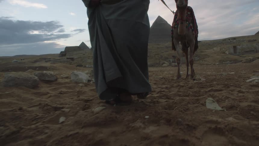 A low-angle, slow-motion view shows an Egyptian man in a traditional galabeya leading a camel across the desert sands with the Pyramids of Giza in the background