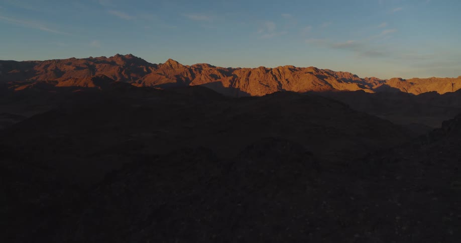 A majestic, wide shot captures the peaks of a rugged desert mountain range illuminated by the warm light of sunrise