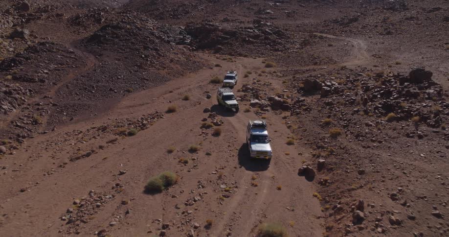 Aerial view of a convoy of 4x4 vehicles driving on a rugged dirt track through a vast, rocky desert mountain range