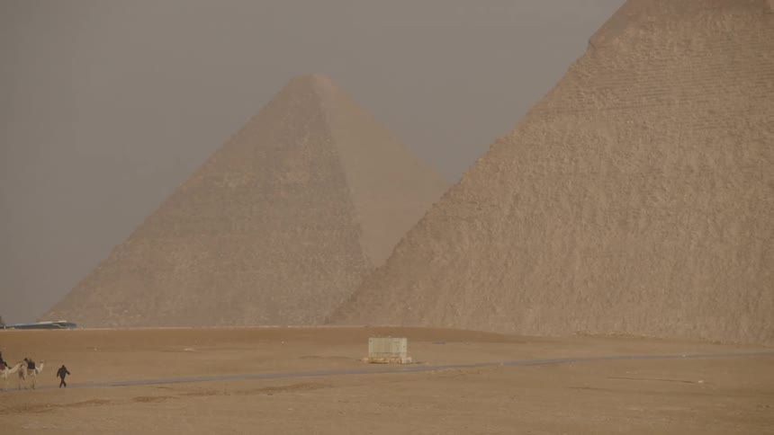 People on a camel travel through the hazy desert with the iconic Giza pyramids looming in the background