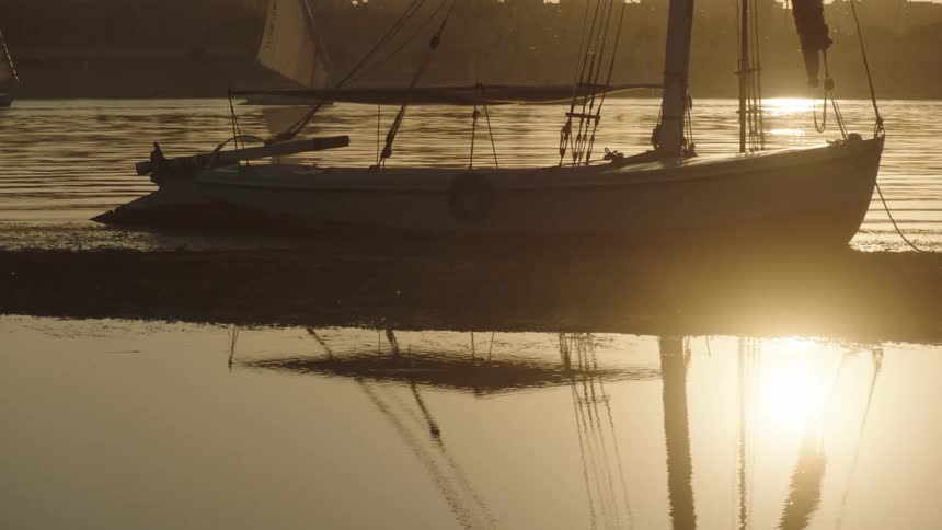 A docked sailboat on the riverbank at golden hour reflects in the calm water as another boat sails in the background