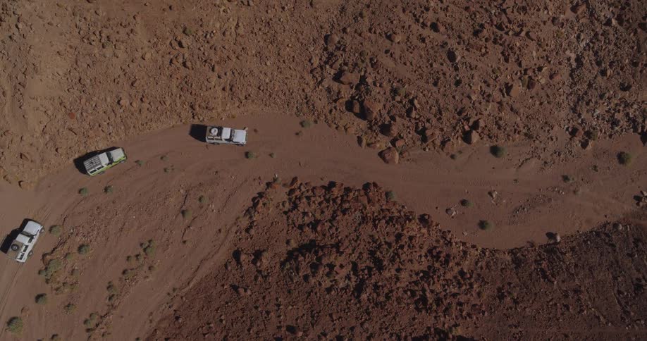 An aerial top-down view shows a convoy of off-road safari jeeps navigating a winding dirt road through a rugged, rocky desert landscape