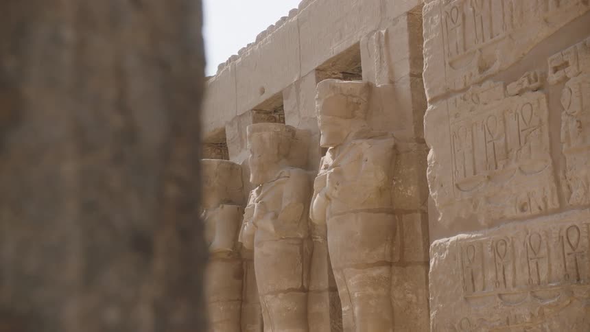 A slow reveal of weathered ancient Egyptian statues and hieroglyphics carved into a sunlit temple wall
