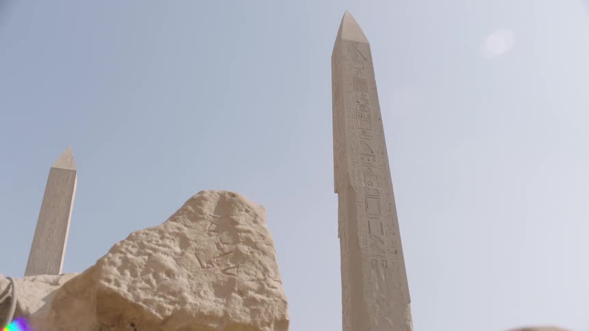 A low-angle pull-back shot reveals ancient Egyptian obelisks with hieroglyphic engravings against a sunny sky