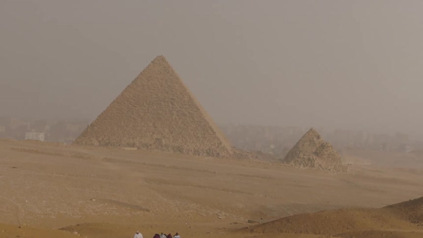A wide shot shows a small group of people walking across the hazy Egyptian desert with the Great Pyramids in the background
