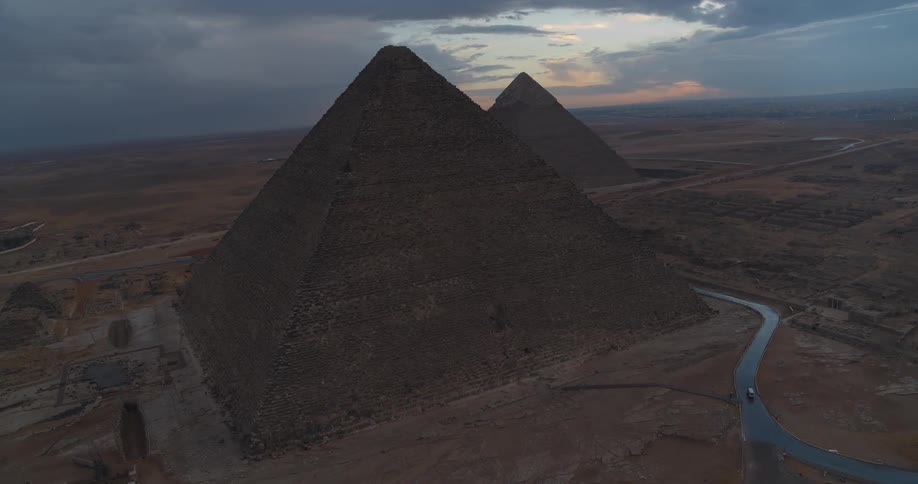 A dramatic aerial view reveals the majestic Pyramids of Giza standing tall in the desert under a cloudy, dark sky at dusk