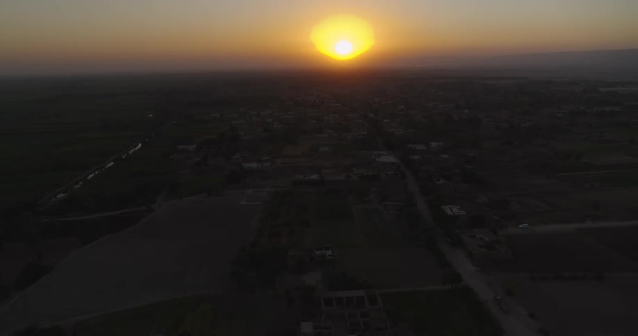 A slow aerial pan over a quiet rural village and surrounding farmland as the sun sets on the horizon