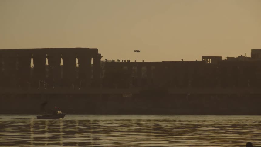 A slow pan across the Nile River at sunset reveals the silhouetted Luxor Temple and a traditional felucca sailboat