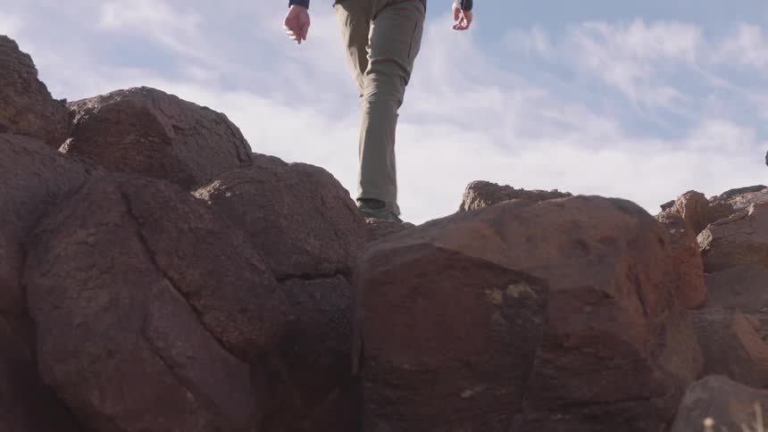 A low-angle slow-motion shot of a hiker's boots carefully navigating and climbing over large rocks against a bright sky