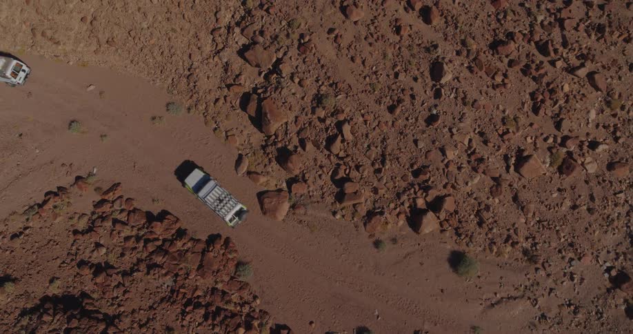 A top-down aerial view of two off-road vehicles navigating a narrow, dusty trail through a remote and rocky desert landscape