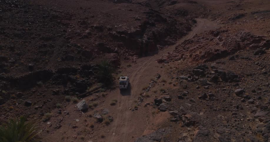 Aerial view of a white 4x4 parked on a dirt track in a rugged, rocky desert canyon as sunlight illuminates the landscape
