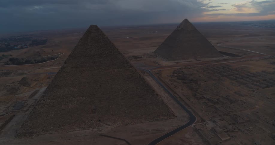 An epic aerial view of the majestic Pyramids of Giza standing silently in the vast desert landscape under a dramatic, cloudy sky at dusk