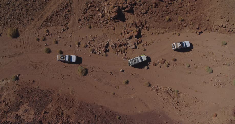 A top-down aerial shot tracks a convoy of three white off-road trucks navigating a dusty, winding trail through a vast and rocky desert landscape
