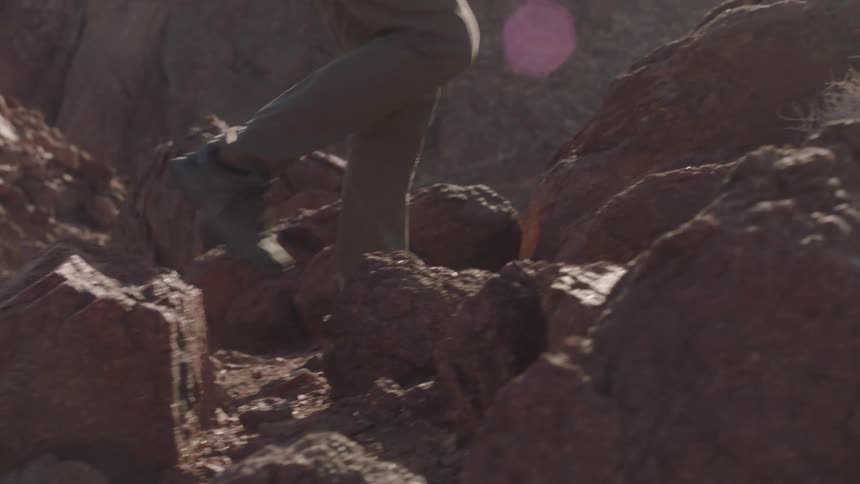 Slow motion close-up of a hiker's boots carefully climbing over a rocky desert trail with a sun flare