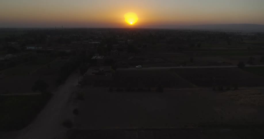 An aerial view shows a lone white car driving down a rural road through farmland as the sun sets on the horizon