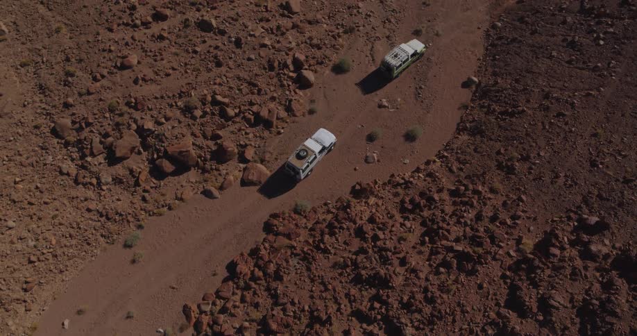 An aerial top-down view shows a convoy of three off-road vehicles driving along a winding dirt road through a remote and rocky desert landscape
