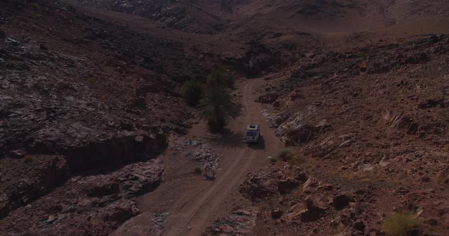 A white off-road vehicle drives on a dusty track through a remote and rocky desert canyon, captured in a slow-pullback aerial shot