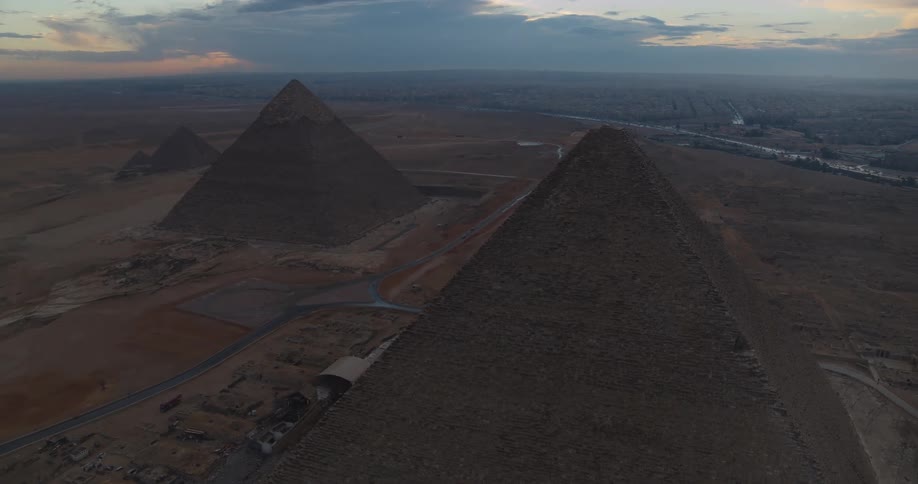 A stunning aerial view rises above the iconic Pyramids of Giza, revealing the ancient structures against the backdrop of a cloudy twilight sky and the distant city