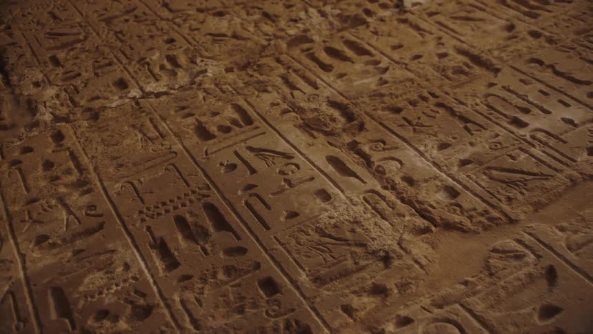 A close-up dolly shot moves across ancient Egyptian hieroglyphics carved into a weathered stone wall