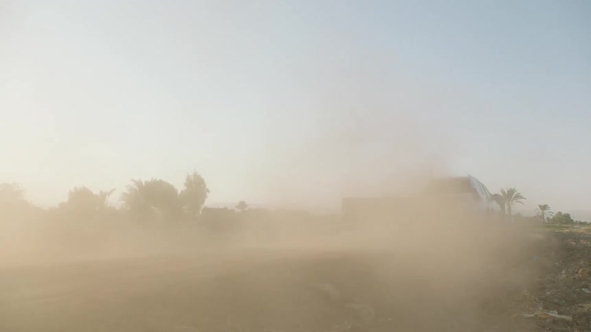 Slow motion shot of a white 4x4 vehicle driving off-road through a dry, rural village, creating a massive dust cloud