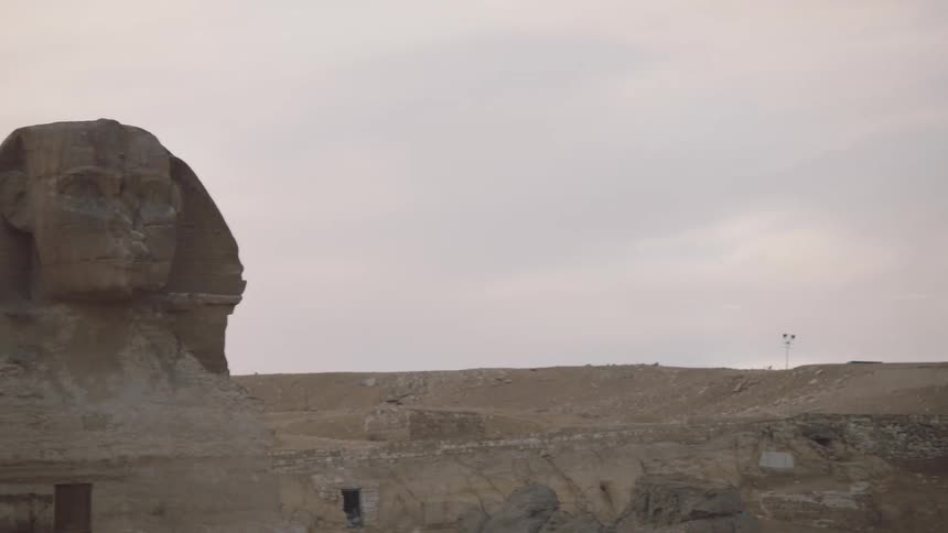 A slow pan reveals the majestic head of the Great Sphinx of Giza with a pyramid in the background under a cloudy sky