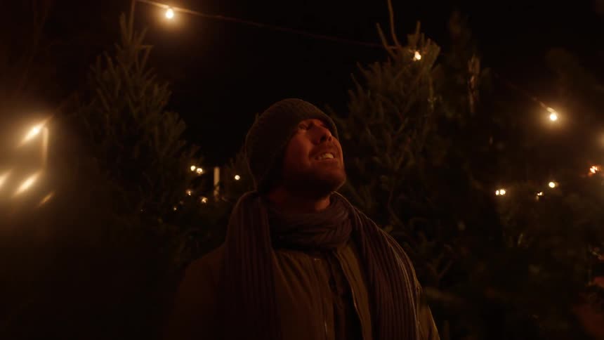 A man standing outside smiling and looking up with  a christmas tree behind him