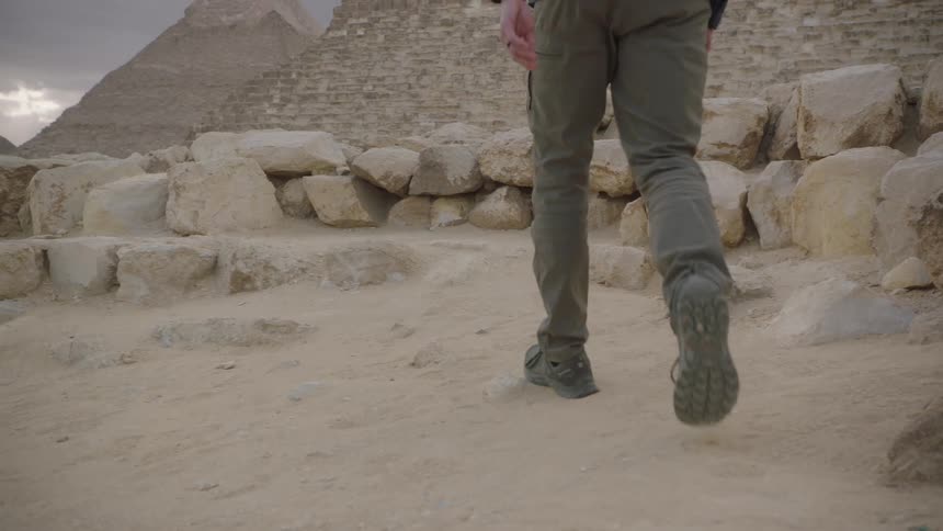 Low angle slow motion shot of a man's legs and feet as he walks through the desert towards the Egyptian pyramids