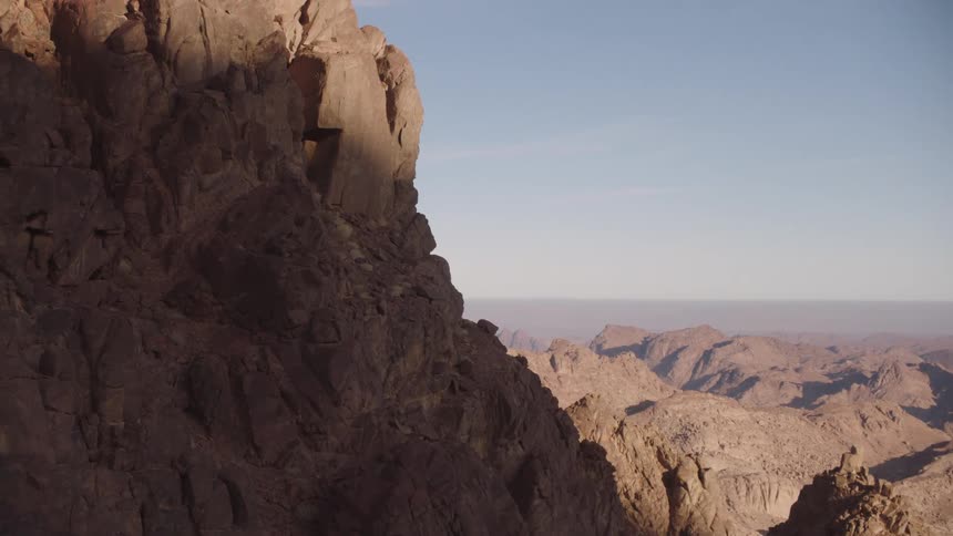 A slow tilt up a shadowed cliff reveals a vast, sun-drenched desert mountain range