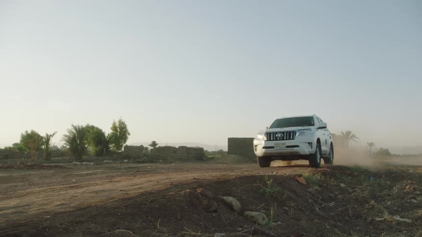 A white SUV drives in slow motion along a dusty dirt road in a remote, arid village, kicking up a trail of dust