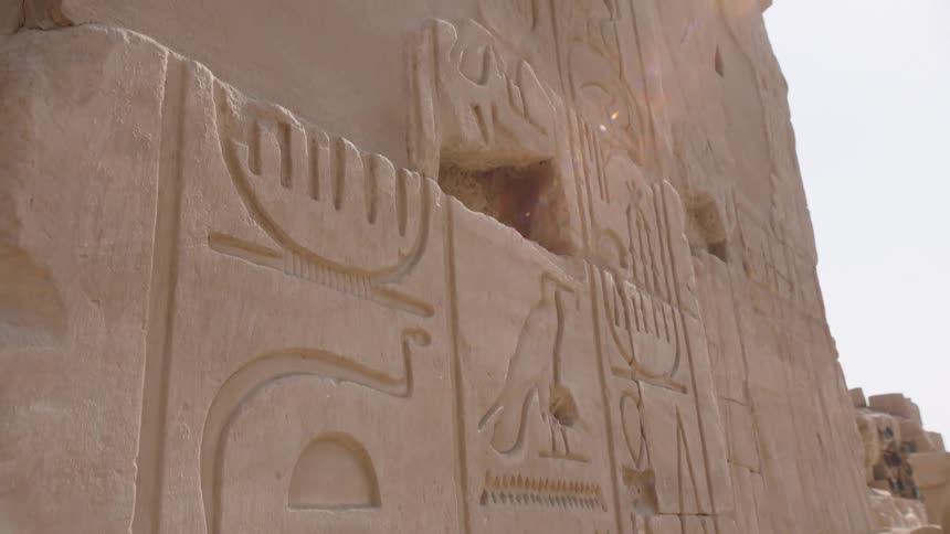 A close-up, upward-tilting shot reveals ancient Egyptian hieroglyphs carved into a stone temple wall as the bright sun creates a beautiful lens flare
