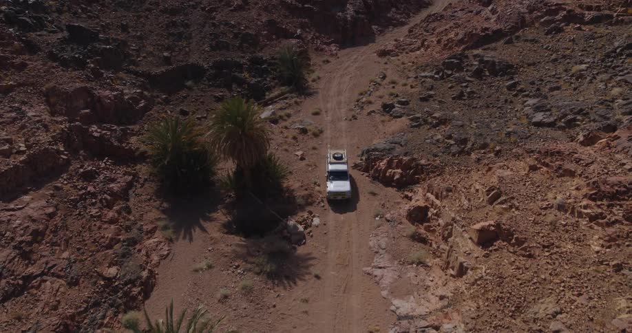 An aerial view shows a white pickup truck driving on a dusty trail through a remote and rocky desert canyon with palm trees