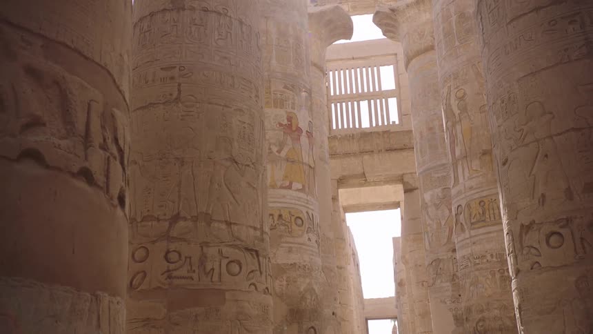 Bright sunlight filters through the massive stone columns of an ancient Egyptian temple, revealing intricate hieroglyphs and faded paintings