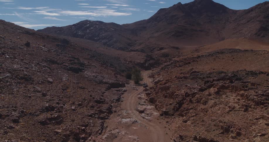 A high-angle aerial shot pulls back from a white SUV on a dirt road to reveal a vast, arid mountain canyon