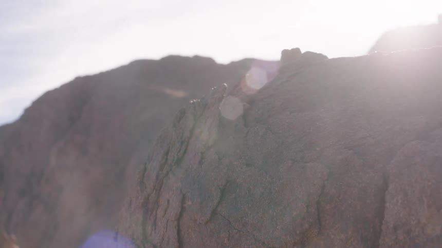 A sun-drenched, static close-up of a large rock formation in a desert landscape with a bright lens flare