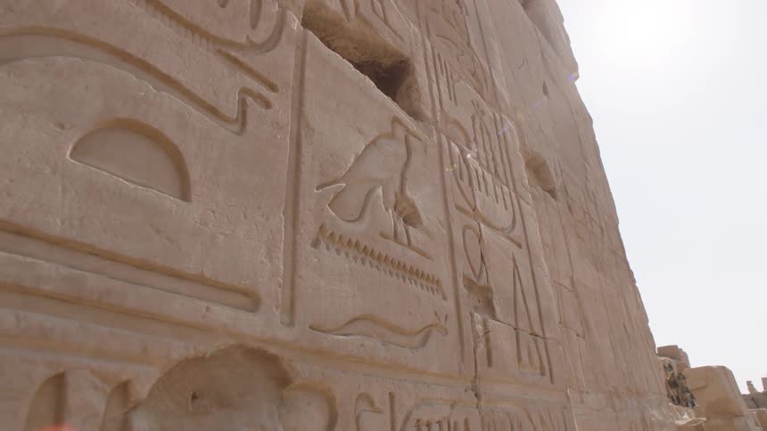 A slow pull-back shot from a low angle reveals detailed ancient Egyptian hieroglyphics carved into a stone wall under the bright sun