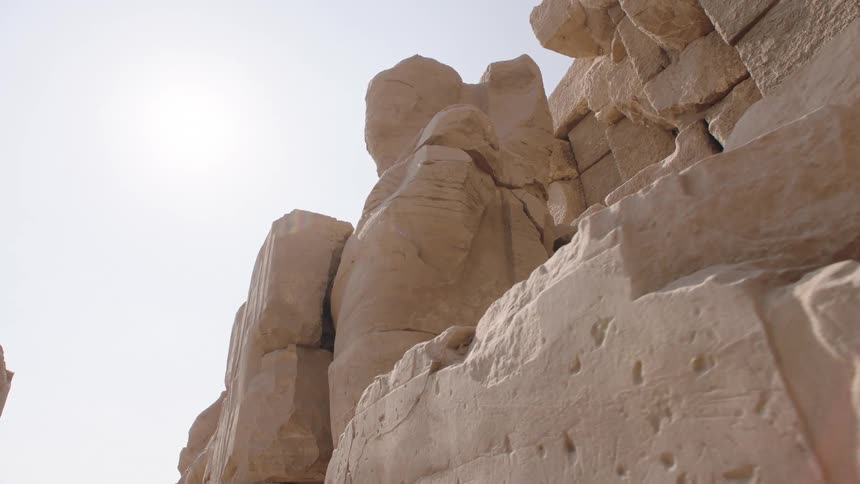 A slow dolly reveals the immense scale of a weathered and crumbling ancient Egyptian statue against the bright desert sun