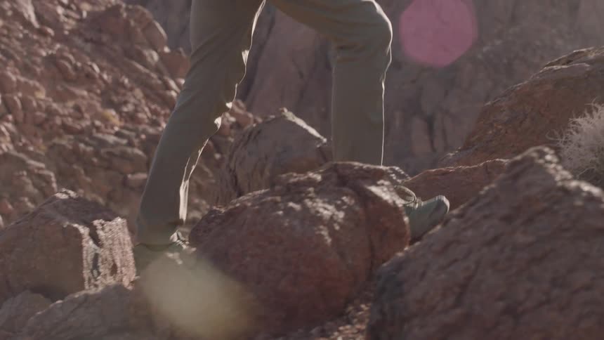 A low-angle slow motion shot shows a hiker's boots carefully climbing up a rocky mountain trail under the bright sun