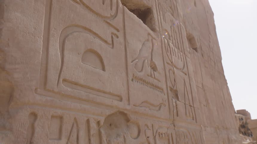 A slow pan down a sunlit stone wall reveals ancient Egyptian hieroglyphic carvings at a historic temple