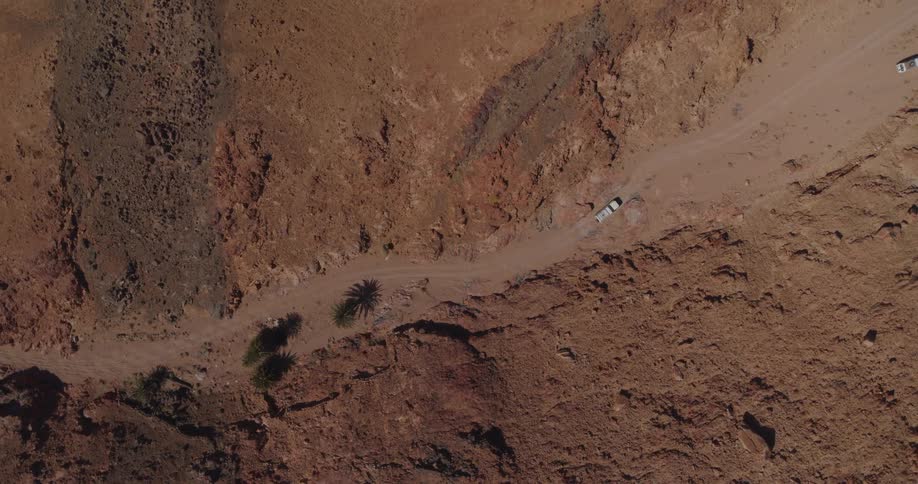 A dramatic aerial top-down shot follows two off-road vehicles as they navigate a winding dirt road through a vast, arid canyon