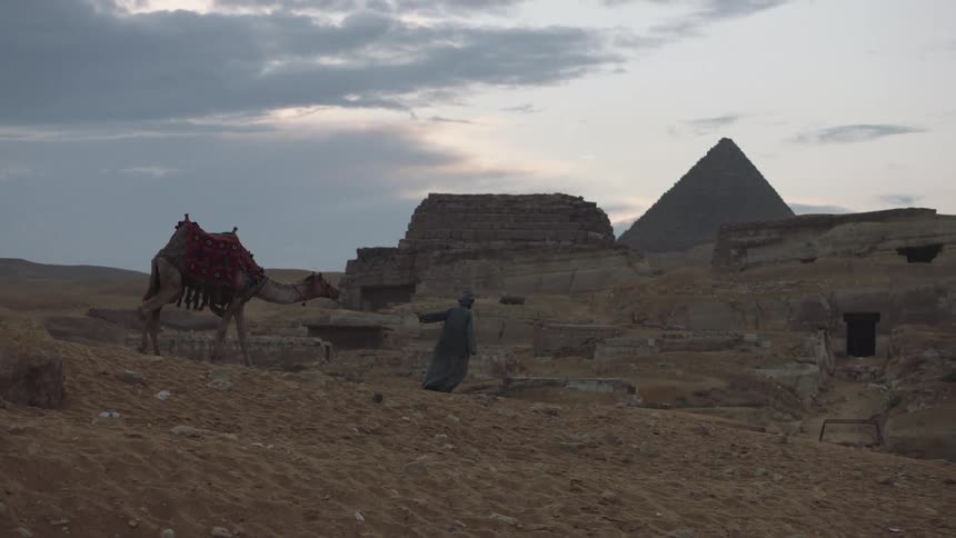 A traditionally dressed Bedouin man leads his camel through the sandy desert landscape with the Great Pyramids of Giza in the background at dusk