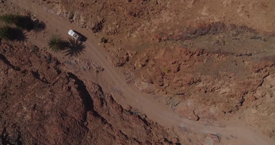 A high-angle top-down aerial shot shows a white off-road vehicle slowly navigating a winding dirt track through a vast and rugged red rock desert canyon