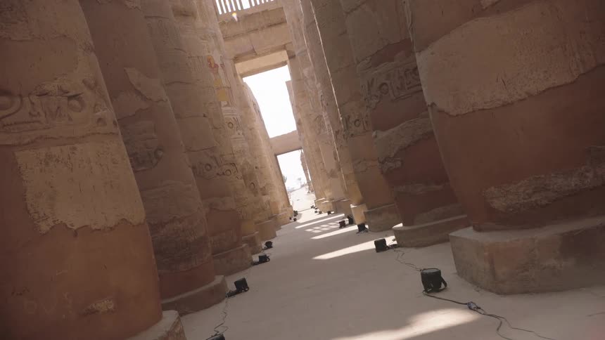 A slow, canted angle shot looks down a corridor lined with the massive, ancient Egyptian columns of the Karnak Temple Complex