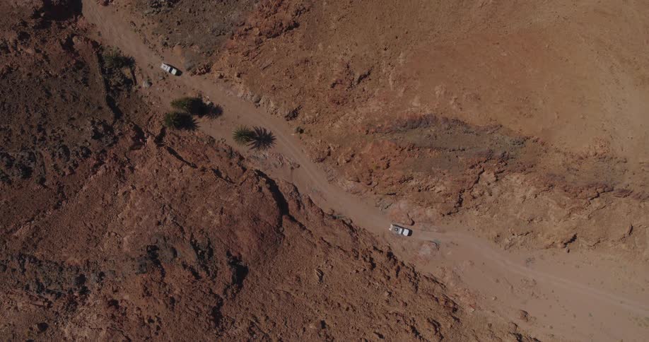 An aerial top-down shot shows a convoy of two white 4x4 vehicles navigating a dusty track through a remote and arid red rock canyon