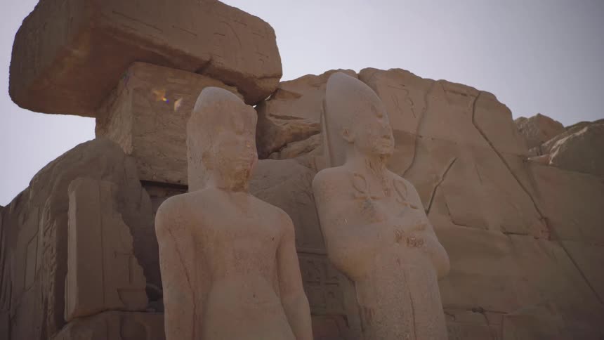Imposing ancient Egyptian pharaoh statues stand amidst temple ruins in a slow-motion dolly shot with sun flare
