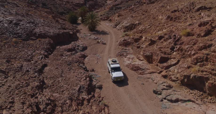 Aerial footage follows a white off-road vehicle as it navigates a winding dirt road through a rugged desert canyon