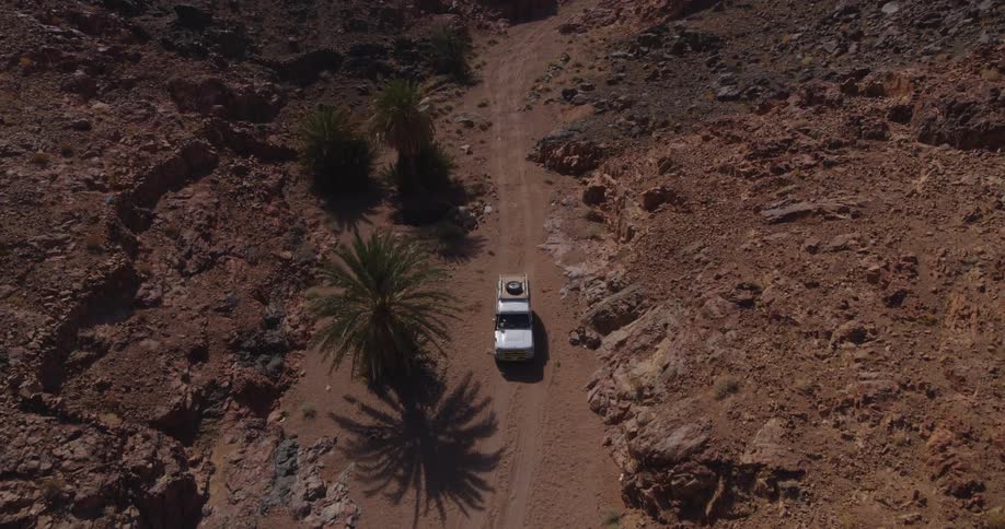 An aerial top-down shot shows a 4x4 vehicle navigating a rugged dirt road through a desolate, rocky desert canyon