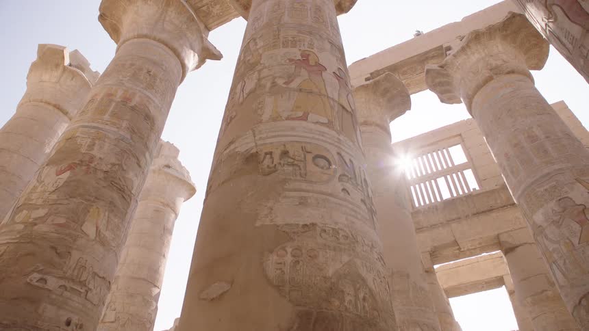 A slow zoom out from towering ancient Egyptian pillars covered in colorful hieroglyphics with the sun flaring brightly between them
